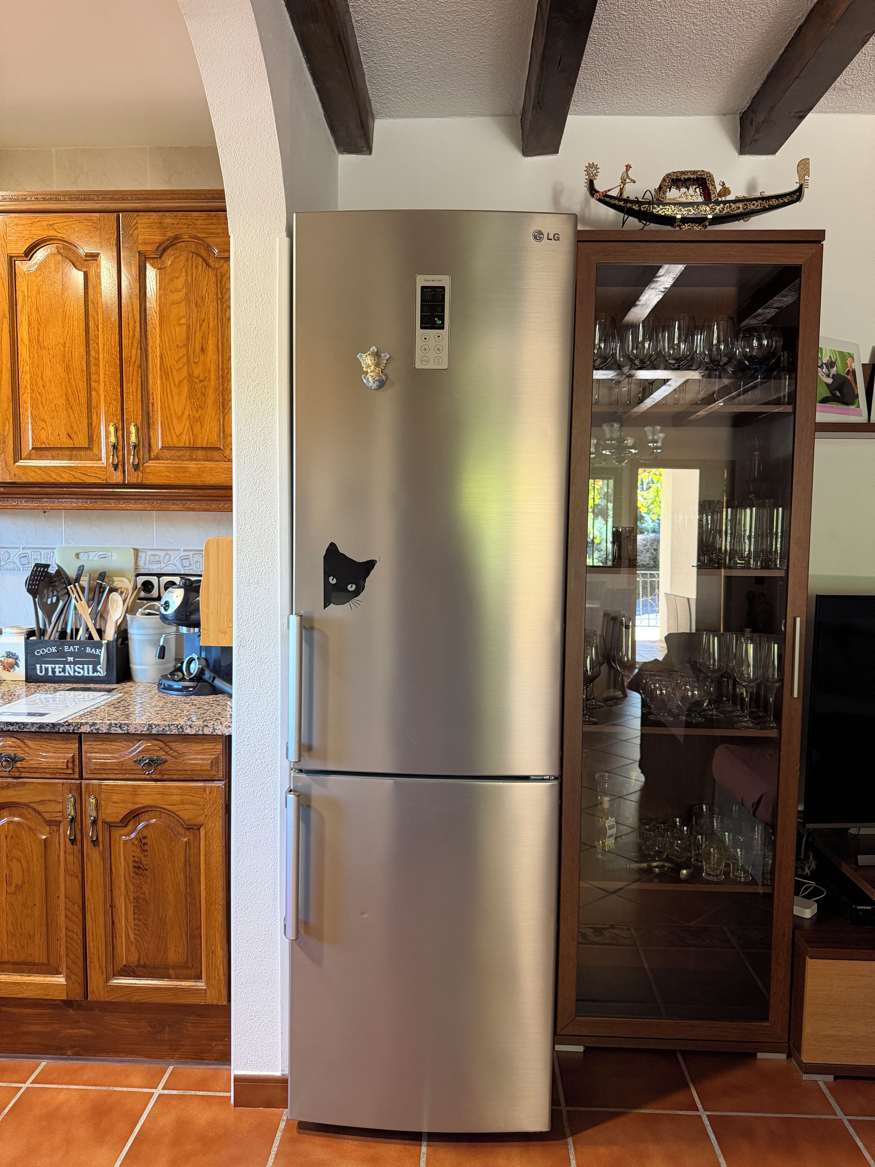 A stainless steel refrigerator with a digital display, wooden cabinets, and a glass-fronted cabinet with wine glasses.