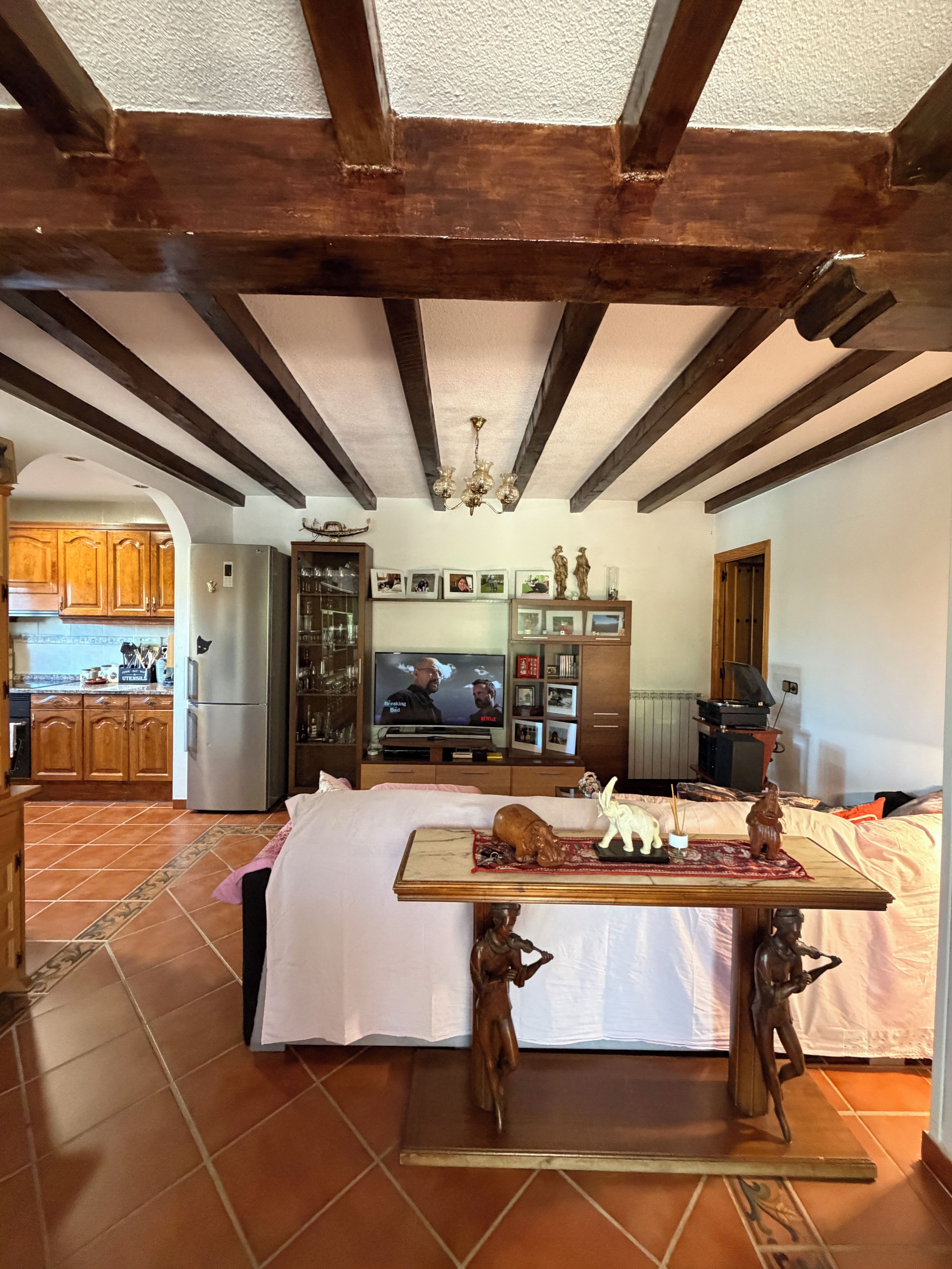 A room with a bed, a table with decorative items, a television, and a kitchen in the background.