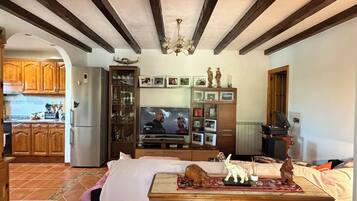 A room with a bed, a table with decorative items, a television, and a kitchen in the background.
