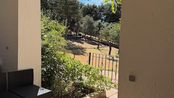 An outdoor patio with a round table and two chairs, a view of a garden with trees and a fence, and a clear sky.
