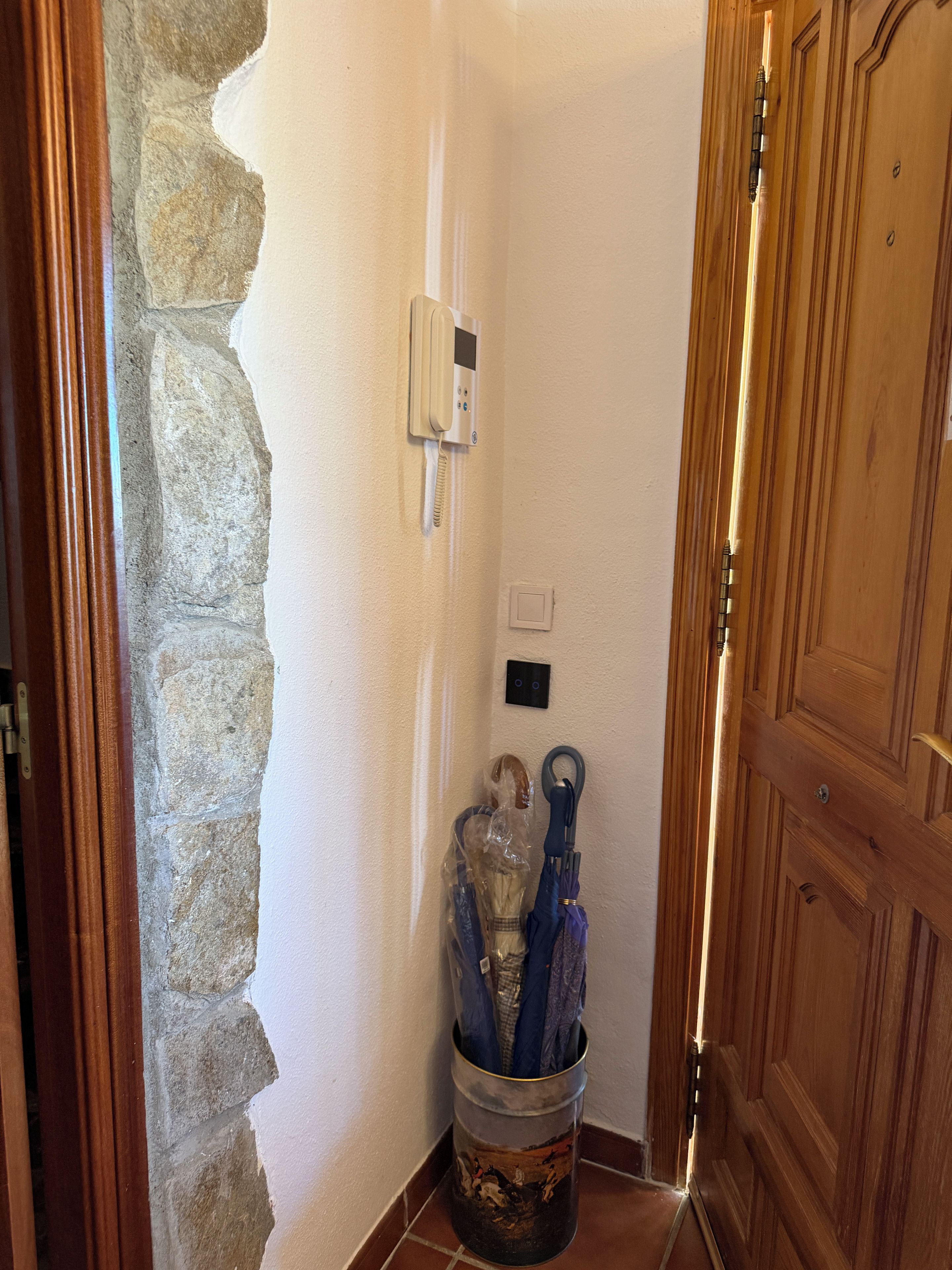 Un pasillo con pared de piedra, puerta de madera, teléfono y cubo de basura con paraguas.