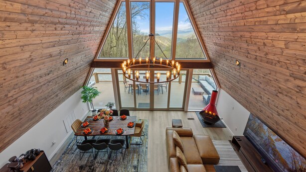 A spacious loft with a high ceiling, wooden walls, a large dining table set for six, a comfortable seating area, and a large window offering a view of the outdoors.