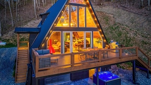 A modern, triangular-shaped cabin with a deck and hot tub, surrounded by a forest.