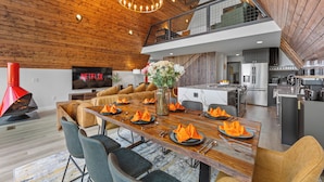 A modern living room with a wooden ceiling, a dining area set for six, a kitchen, and a TV displaying 'NETFLIX'.