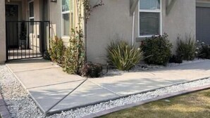 Une maison à deux étages avec un porche à l’avant, un petit jardin avec des plantes et un ciel dégagé.