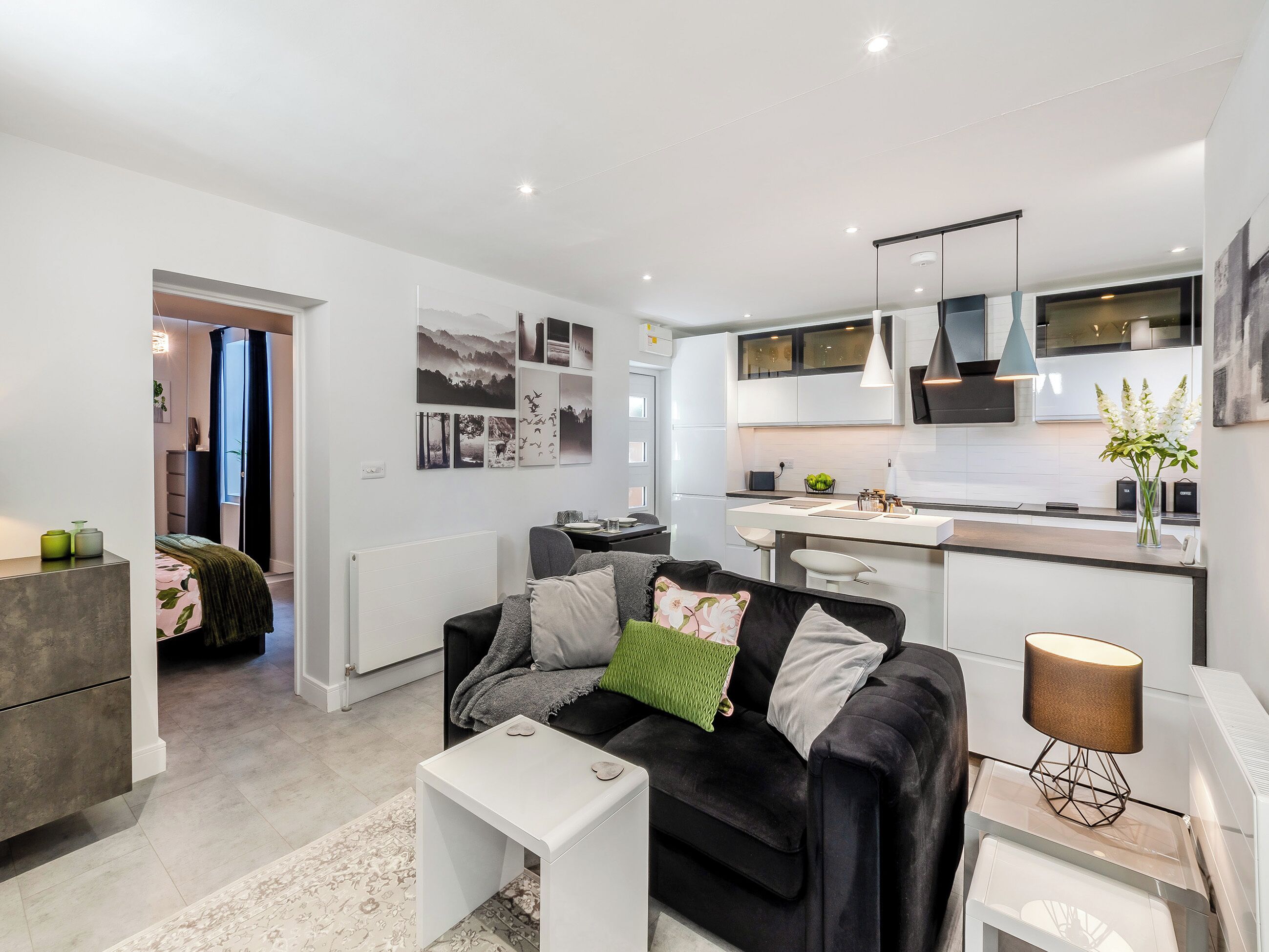 Een moderne woonkamer met een zwarte bank, een witte salontafel en een bijzettafeltje met een lamp. Achtergrond toont een keuken met witte kasten en een centraal eiland.