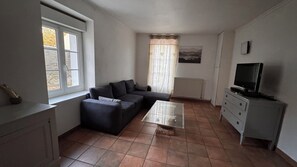 Un salon comprenant un canapé d’angle, une table basse en verre, un téléviseur posé sur un meuble, une fenêtre avec des rideaux et un radiateur.