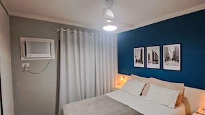 A bedroom with a bed, white curtains, a ceiling fan, a wall-mounted air conditioner, and three framed pictures on the wall.