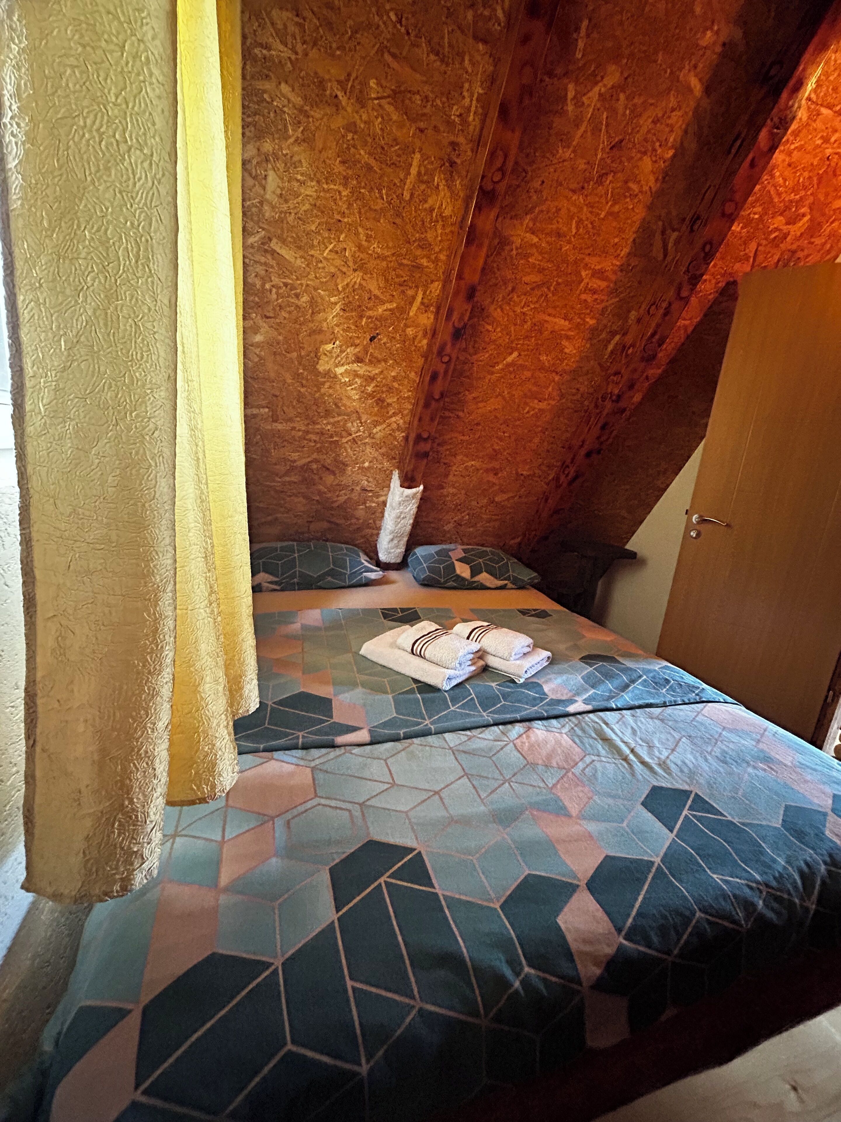 Une chambre avec un plafond en pente, un lit recouvert d’une couvre-lit à motifs géométriques, deux oreillers et une paire de serviettes pliées posées sur le lit.