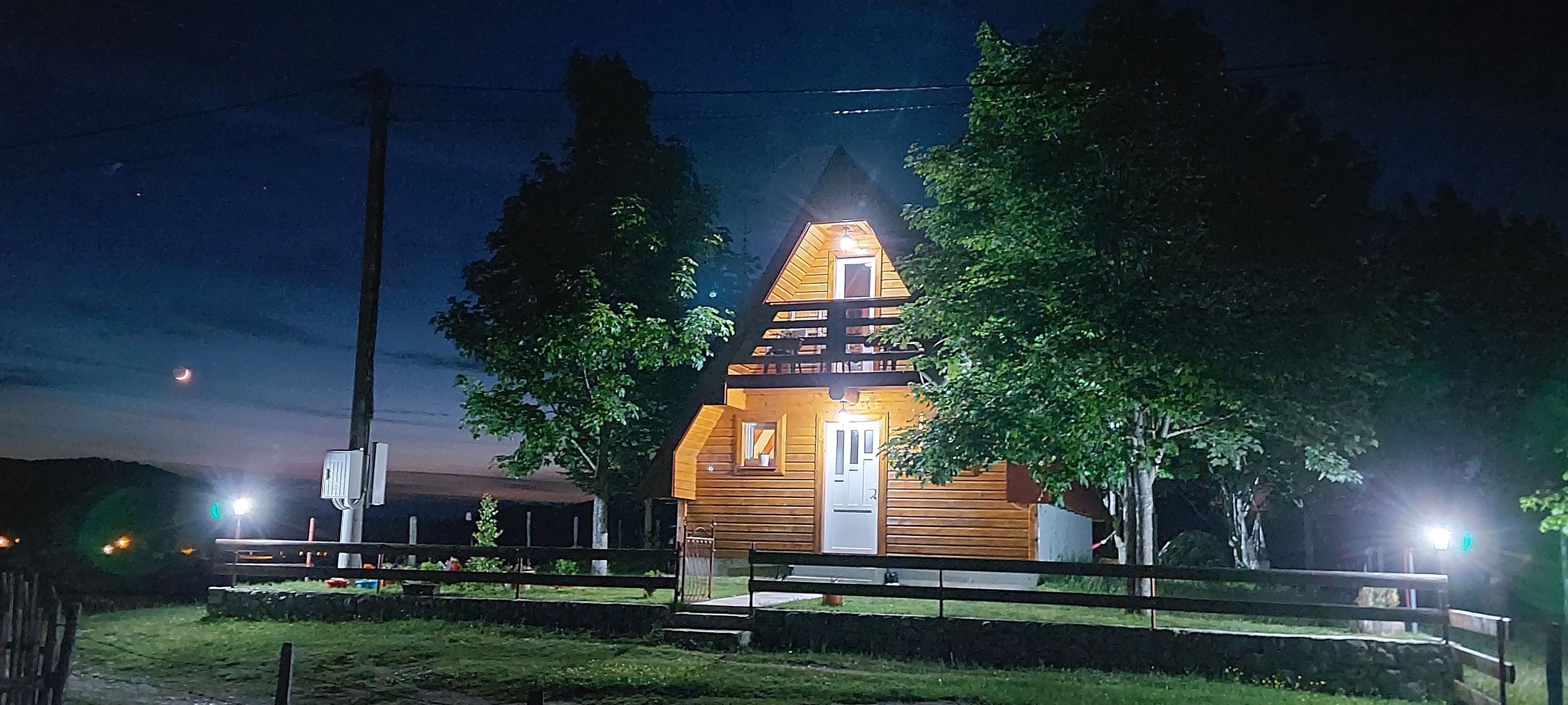 A small wooden cabin with a sloped roof, surrounded by a wooden fence, and illuminated by exterior lights.