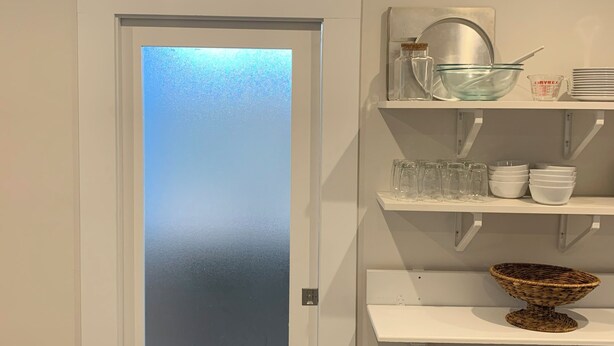Ein Zimmer mit einer Tür aus mattiertem Glas, weißen Regalen mit Gläsern und einem gewebten Korb.
