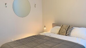 Ein ordentlich bezogenes Bett mit grauem Duvet, ein runder Spiegel an der Wand und eine Nachttischlampe.