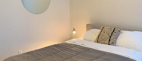 A neatly made bed with a grey duvet, a round mirror on the wall, and a bedside lamp.