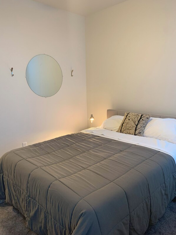 Una cama bien hecha con edredón gris, un espejo redondo en la pared y una lámpara de noche.