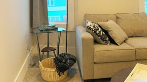 Ein Wohnzimmer mit einer Couch, einem kleinen Beistelltisch, einer Stehlampe, einem Korb und einem Fenster mit Vorhängen.