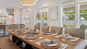 A dining area with a long wooden table set for six, surrounded by chairs, with a chandelier above and a view of the outdoors through large windows. - Luxe Beach Retreat Walk to Beach Pool Spa, Golf Cart, Game Room & Putting Green (Santa Rosa Beach)