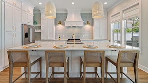 A modern kitchen with white cabinetry, a central island with bar stools, pendant lighting, and a view to the outside. - Luxe Beach Retreat Walk to Beach Pool Spa, Golf Cart, Game Room & Putting Green (Santa Rosa Beach)