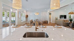 A modern kitchen with a large island, two hanging wicker chandeliers, and a dining area with a wooden table and chairs. - Luxe Beach Retreat Walk to Beach Pool Spa, Golf Cart, Game Room & Putting Green (Santa Rosa Beach)