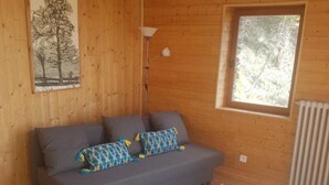 A cozy room with a wooden interior, a gray sofa with patterned cushions, a blue rug, and a large window with a view of trees.