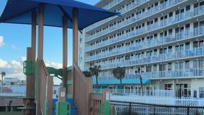 A playground with a blue canopy and wooden play structures, a blue barrel, and a multi-story hotel building in the background. - Gorgeous Views/Beautiful Oceanfront Condo, Beach, Pool/Hot Tub, 4th Fl, 2-Bath (Daytona Beach)