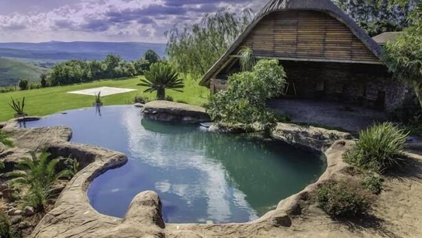 Una casa de piedra rústica con techo de paja, rodeada por un paisaje natural y un pequeño estanque.