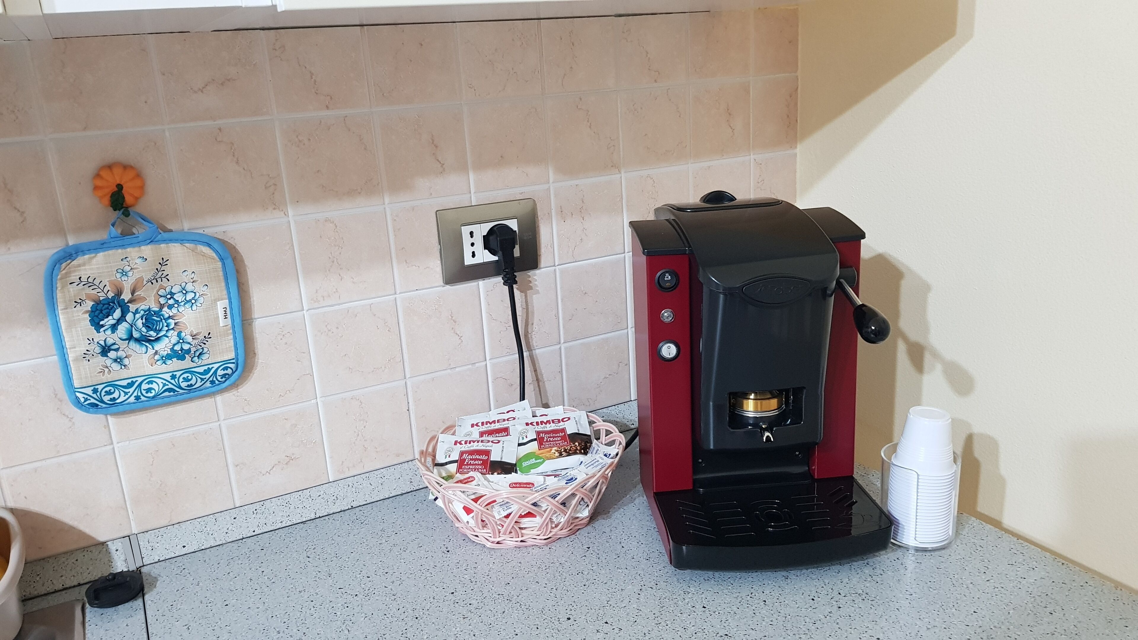 A kitchen with a coffee machine, a basket of coffee pods, and a blue floral kitchen towel on the wall.