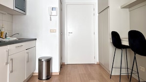 A compact kitchen with white cabinets, a stainless steel trash bin, a microwave, and a small black chair. - Studio Apartment Heart of Brussels (Brussels)