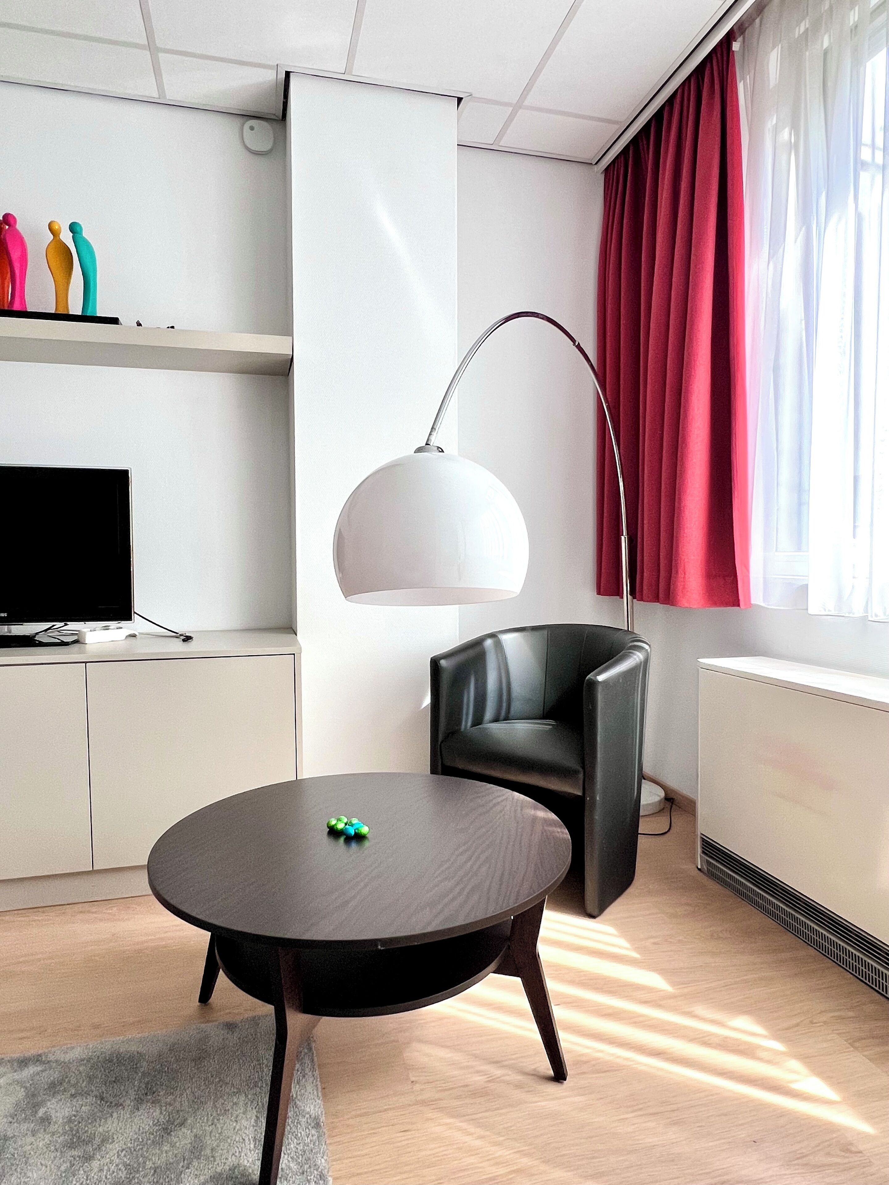 Una sala de estar moderna con una mesa redonda, un sillón de cuero negro, una lámpara de pie, un televisor de pantalla plana y un radiador.