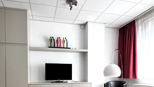 A modern hotel room with a bed, a flat-screen TV, a desk with a lamp, and a window with red curtains. - Studio Apartment Heart of Brussels (Brussels)