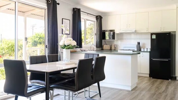 A modern kitchen with a central island, black appliances, and a dining area with black chairs. - Lake Cove Daylesford (Daylesford)