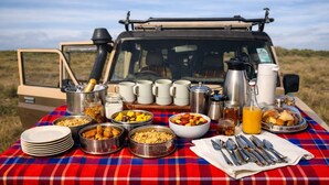 An outdoor dining setup with a variety of food items and utensils on a checkered tablecloth, with a vehicle in the background. - Kapoto Camp (Maasai Mara)