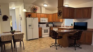A kitchen with wooden cabinets, a refrigerator, a microwave, an oven, a dining table with chairs, and a bar area with stools.