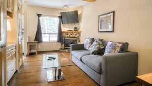 Ein gemütliches Wohnzimmer mit grauem Sofa, einem Glastisch, einem Kamin und einem an der Wand befestigten Fernseher.
