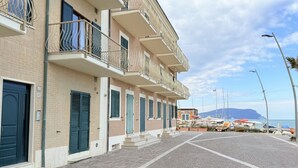 Una hilera de edificios de varios pisos con balcones, una calle empedrada y barcos al fondo.