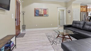 A living room with a leather sofa, a glass coffee table, and a TV mounted on the wall. There is a door leading to another room and a coat rack near the door. - Charming 3-bedroom house located minutes from LSU (Baton Rouge)