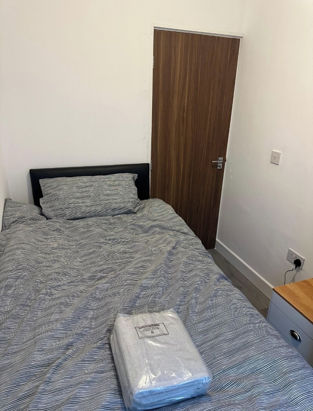 A small, neatly arranged bedroom with a single bed, a wooden wardrobe, and a small bedside table.