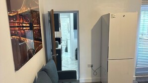 A modern living room with a grey sofa, a wooden coffee table, and a white refrigerator. A large cityscape painting is on the wall, and there is a doorway leading to a kitchen area.