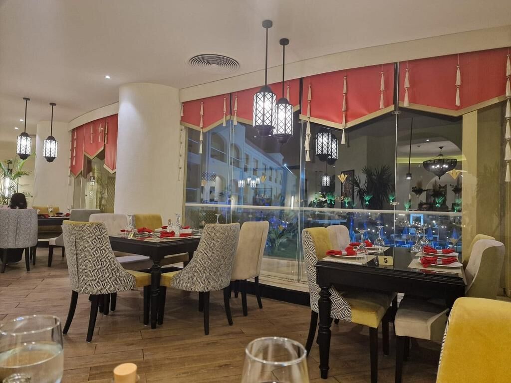 A restaurant with multiple dining tables and chairs, large windows with a view of the city, and hanging pendant lights.