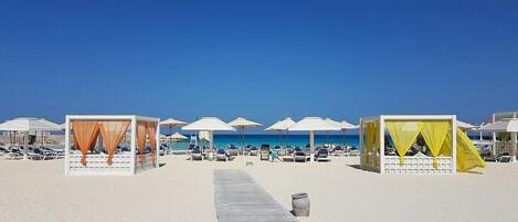 A beach with white beach umbrellas and sun loungers, a wooden walkway, and a clear blue sky.