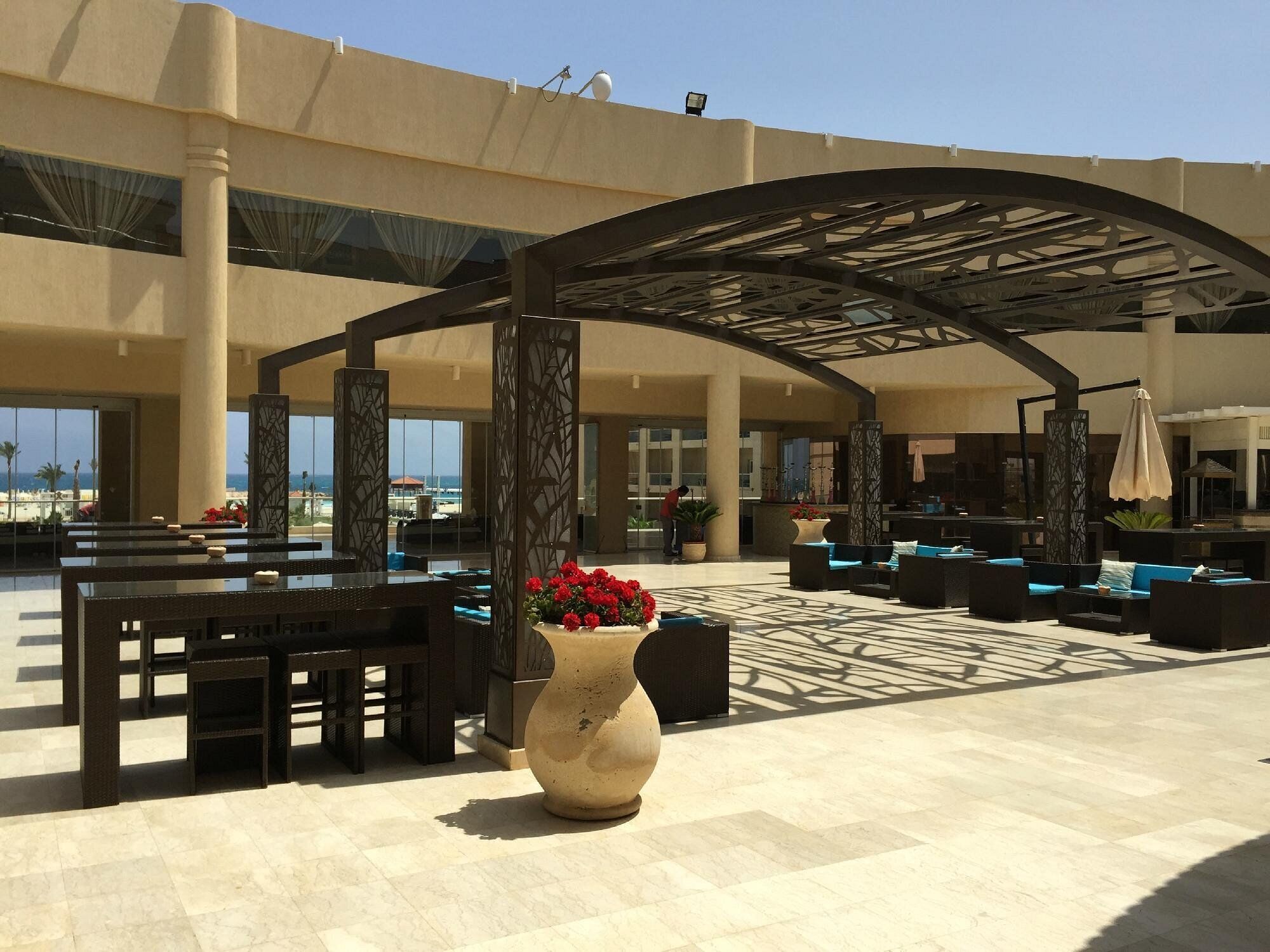 Un área de estar al aire libre con mesas y sillas, un jarrón decorativo grande con flores, y una estructura de pérgola.