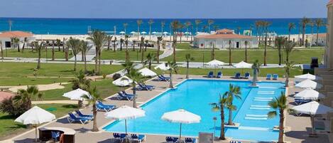 A swimming pool with lounge chairs and umbrellas, surrounded by palm trees and a view of the ocean.