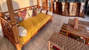 Un lit traditionnel en bois, agrémenté d’oreillers et d’une couvre-lit jaune, un banc en osier, un tapis rayé aux couleurs vives et un mur de pierre avec une fenêtre donnant sur la verdure.