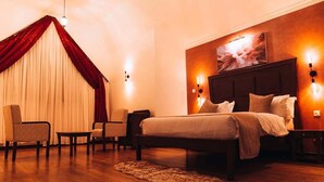 Un dormitorio con una cama grande, dos mesitas de noche, cabecero con apliques de pared, un ventanal con cortinas y suelo de madera.