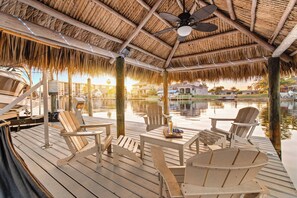 A covered deck with wooden chairs and tables, a boat docked nearby, and a calm water body in the background. - Fall Szn Sale! Luxe Villa with Mini Golf, Kayaks, Hot Tub, Game Room & Karaoke (Cape Coral)
