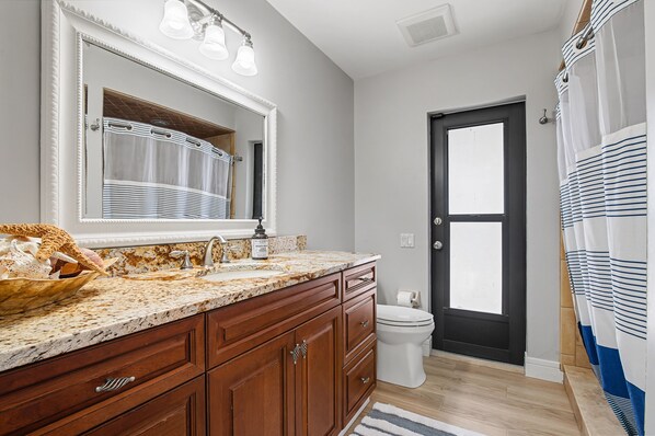 A bathroom with a large mirror, a sink with a marble countertop, wooden cabinets, a toilet, and a shower curtain. - Fall Szn Sale! Luxe Villa with Mini Golf, Kayaks, Hot Tub, Game Room & Karaoke (Cape Coral)