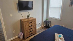 Un dormitorio con una cama, un aparador de madera, un televisor montado en la pared, una ventana con persianas y una pequeña mesa con una planta.