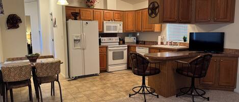 A kitchen with wooden cabinets, a refrigerator, a microwave, an oven, a dining table with chairs, and a bar area with stools.