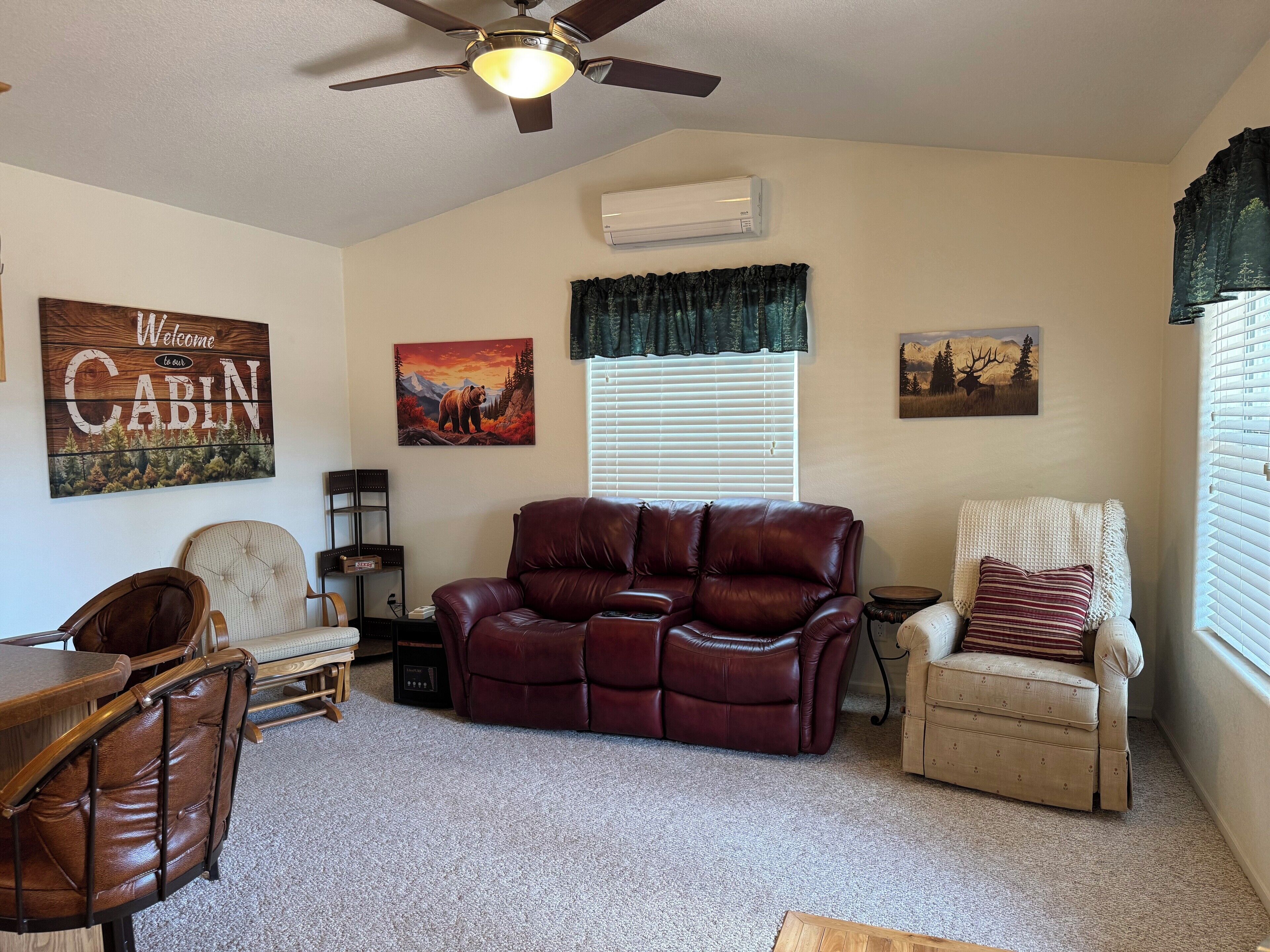 A living room with a leather sofa, a wooden coffee table, a recliner, and a television. There is a ceiling fan, a painting of a bear, and a sign that says "Welcome to our cabin."