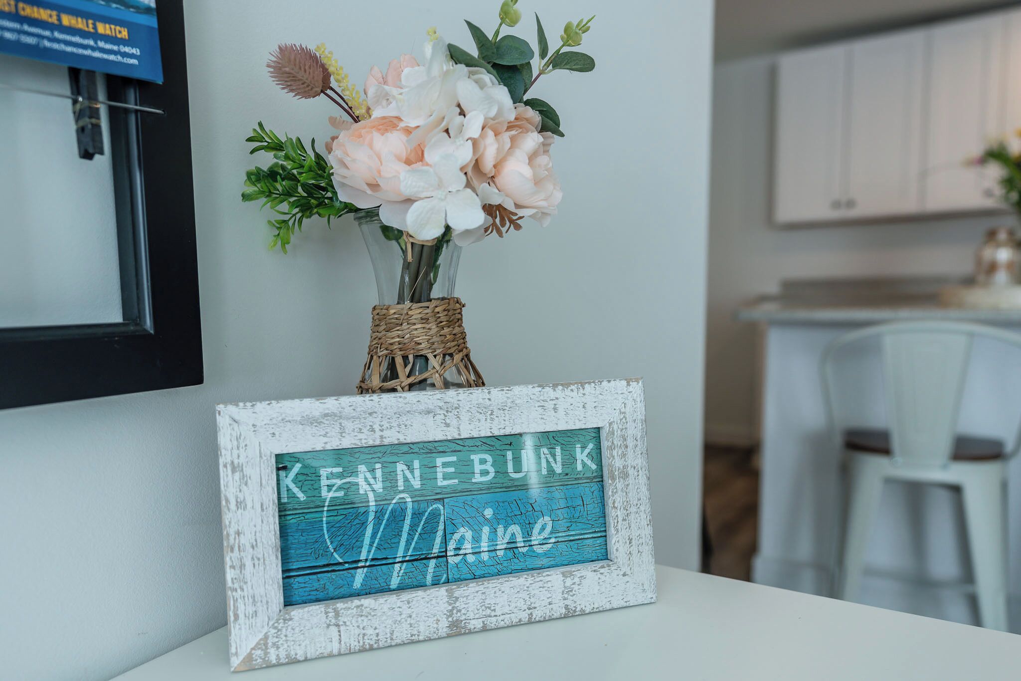 Un coin cuisine avec des meubles blancs, un vase en osier rempli de fleurs et une pancarte sur laquelle on peut lire « KENNEBUNK, Maine ».