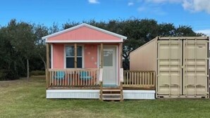 A small pink cabin with a wooden deck and a beige shipping container attached to its side. - Custom built cool cabin with AC in Holiday Beach. Not a converted shed. (Rockport)